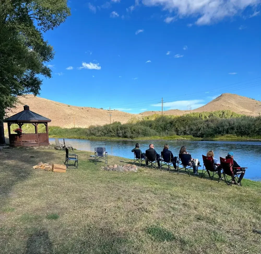 Guests relaxing in camp chairs beside the Rio Grande, with a wooden gazebo, shade trees and hills beyond.