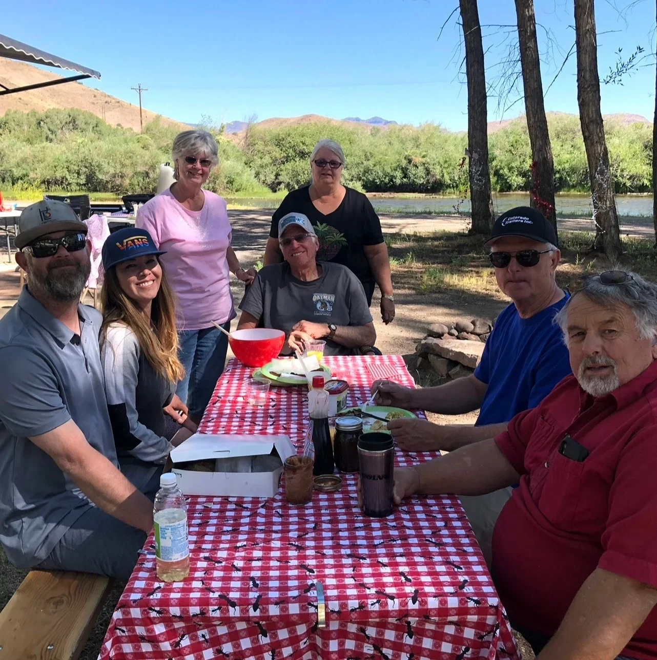 Guests gathered around a picnic table beside the Rio Grande beneath shade trees.