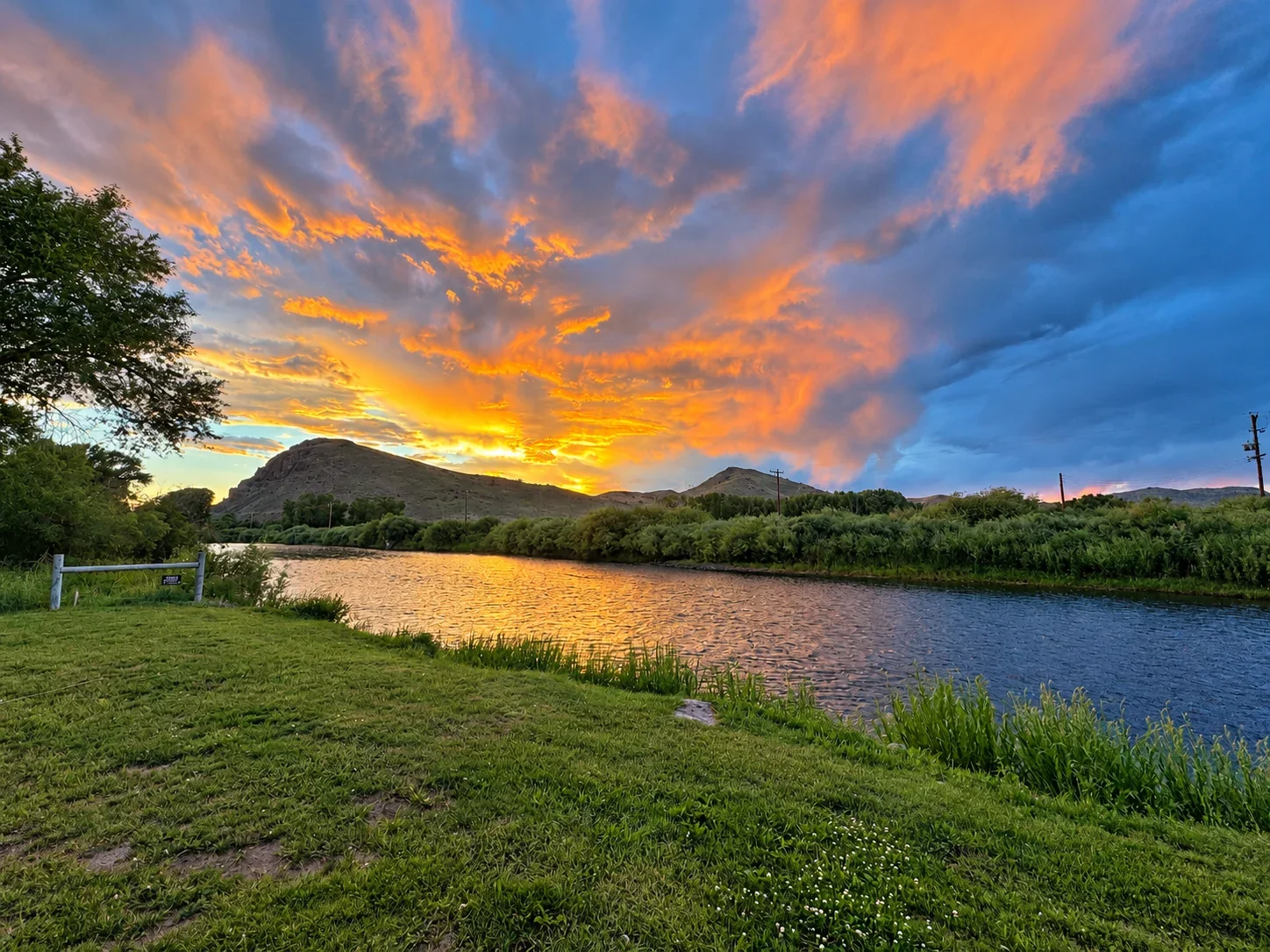 Orange sunset clouds reflected in the Rio Grande beside a grassy riverbank.