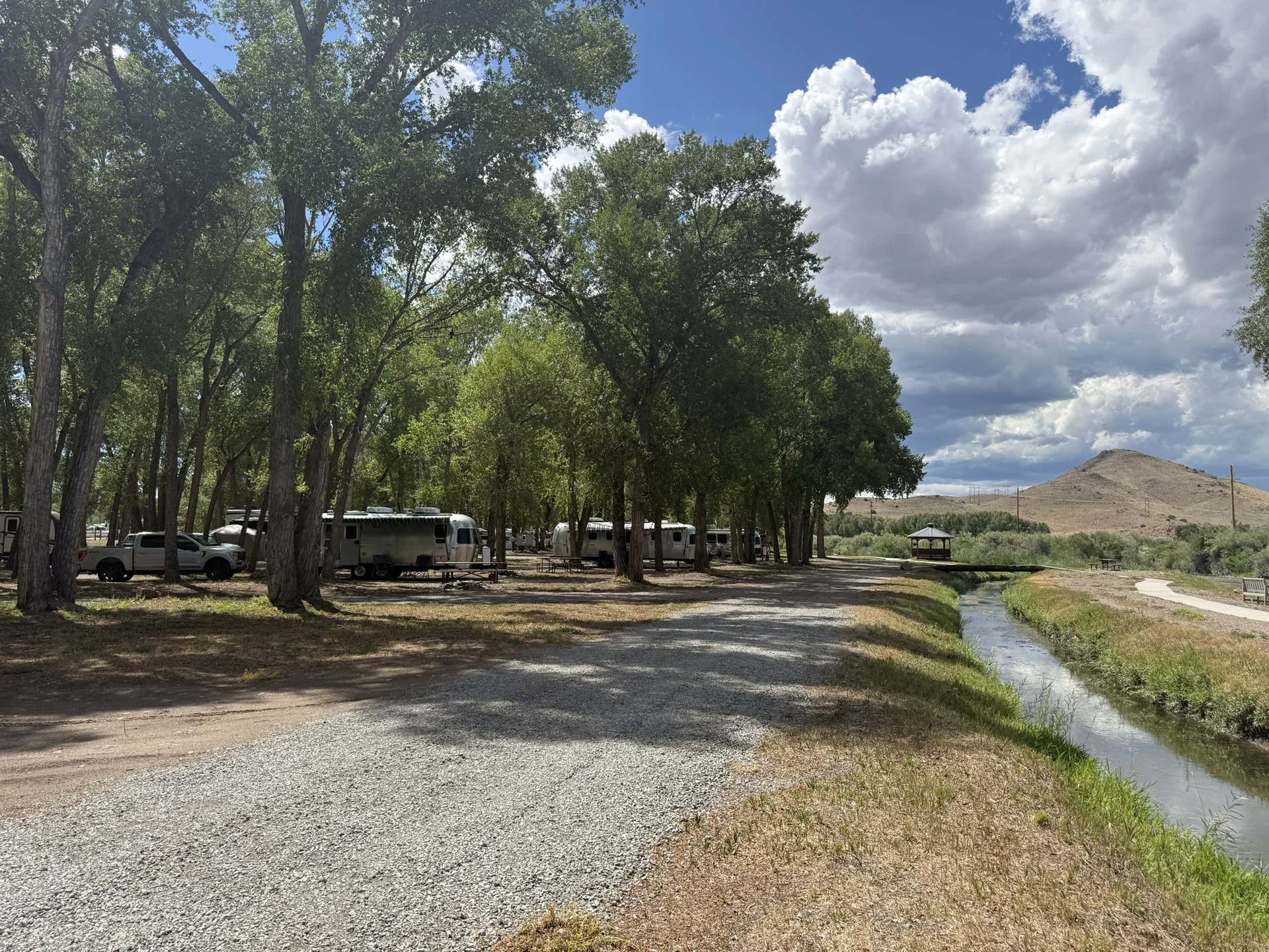 Tree-shaded RV sites beside a broad gravel lane, a narrow water channel and a gazebo.