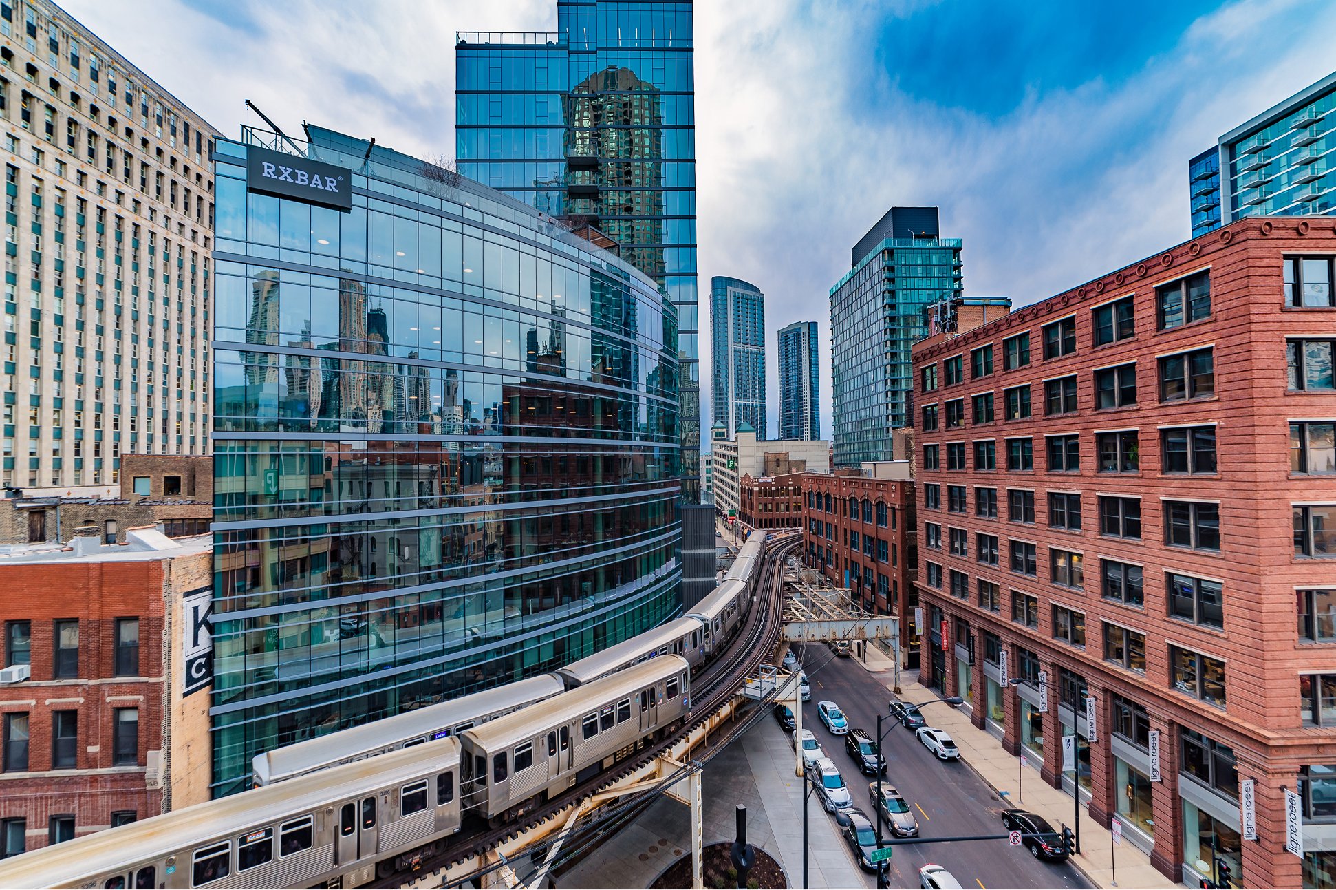 A train moves along a city street, flanked by tall buildings in the background, showcasing urban life and transportation.