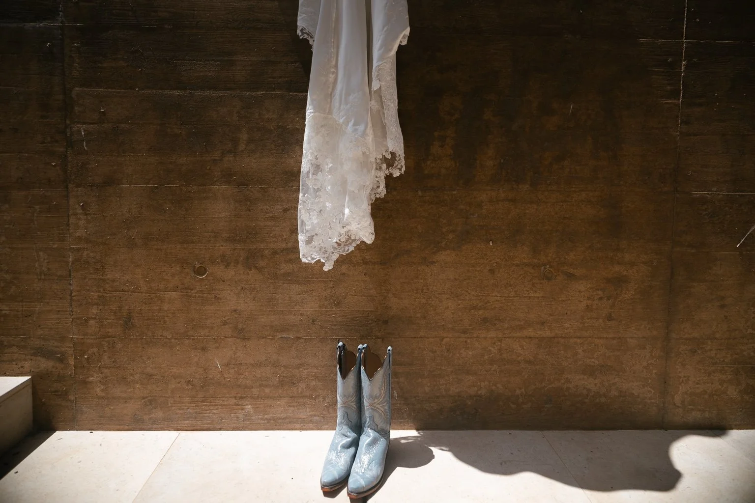 Lace wedding dress hanging above pale blue cowboy boots against a concrete wall.