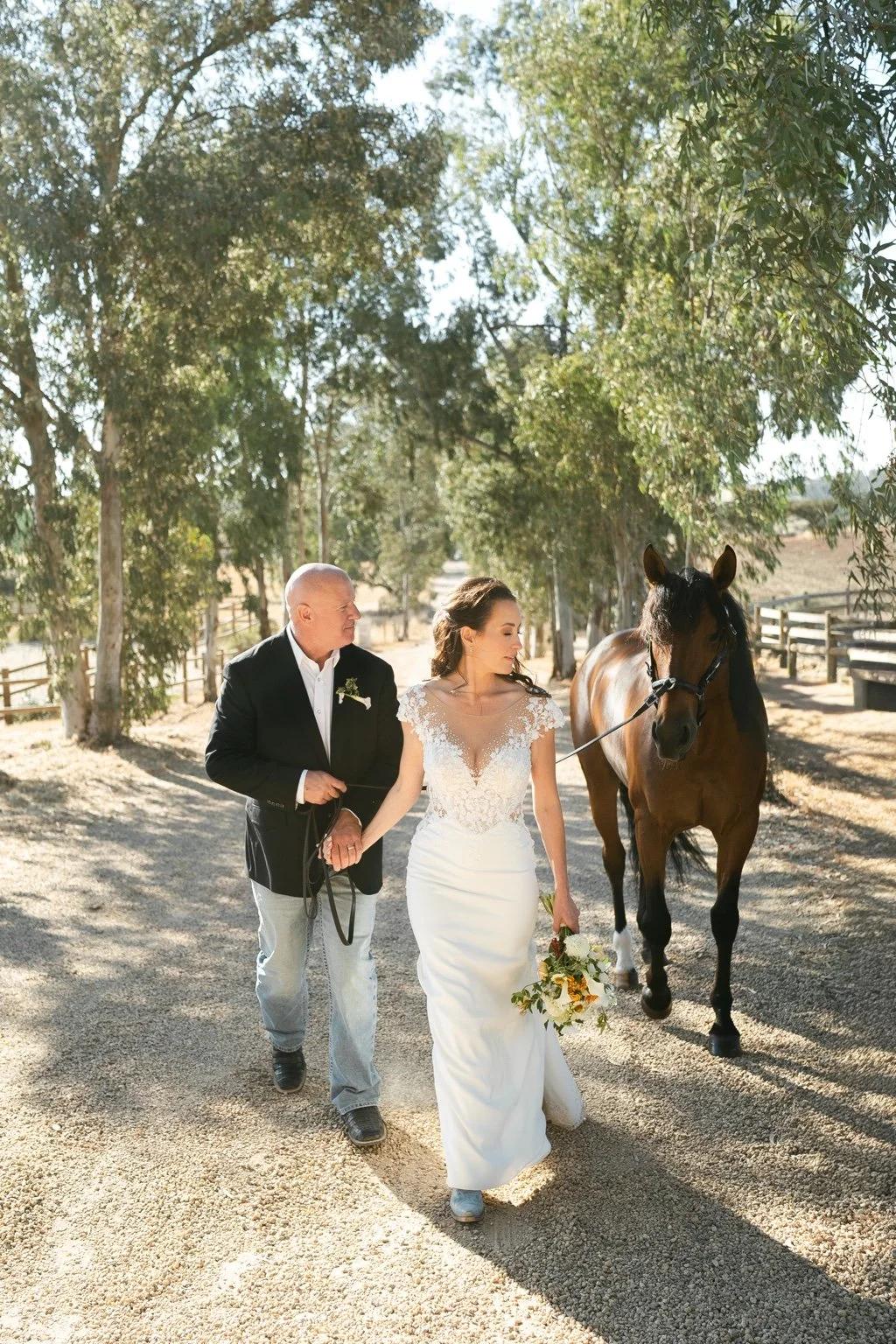 Joanna and Greg walking beside a brown horse along a tree-lined path.
