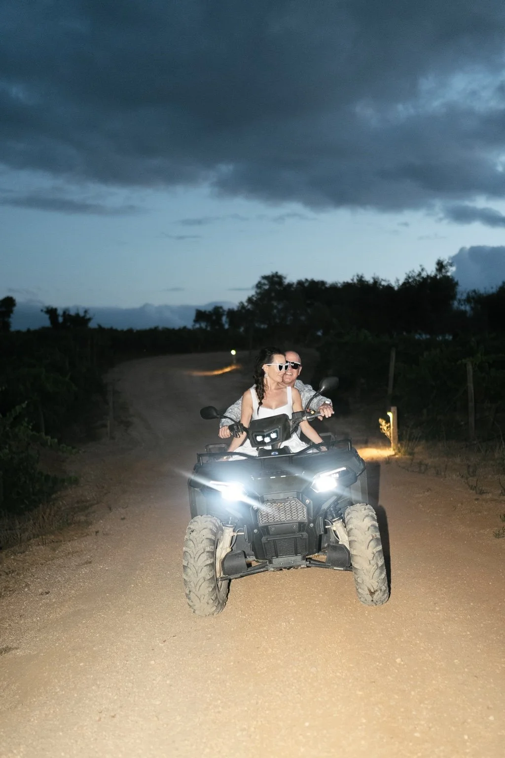 Joanna and Greg riding a quad bike on a rural path at dusk.