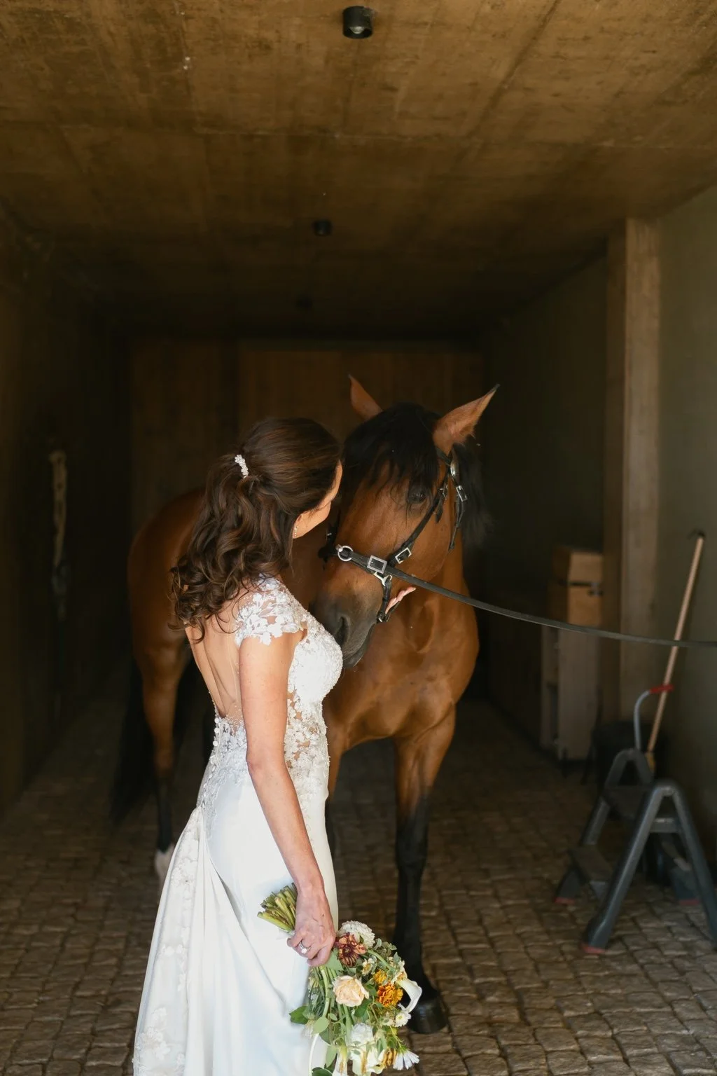 Joanna beside a brown horse inside a shaded stable.