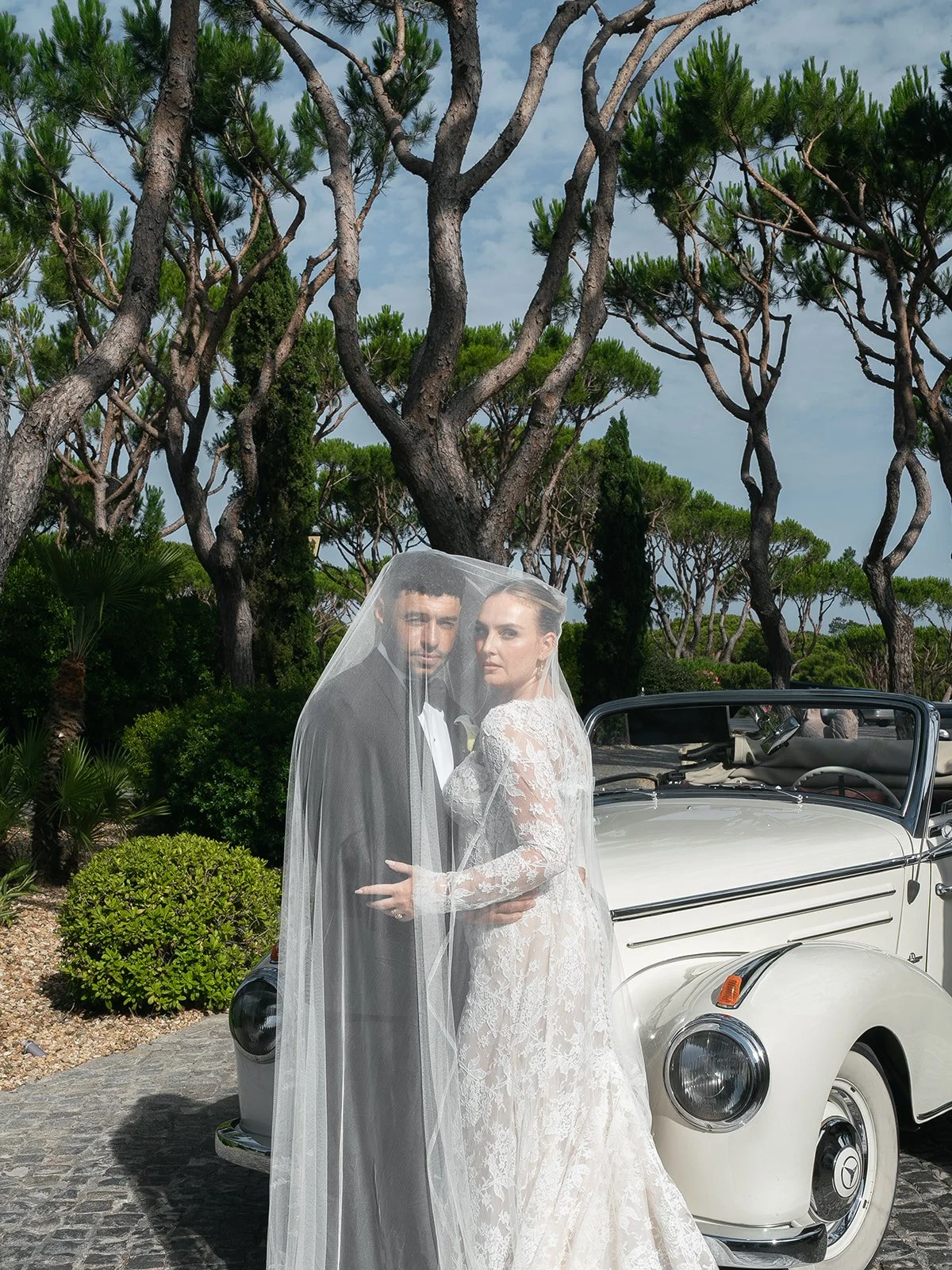 Perrie Edwards and Alex Oxlade-Chamberlain beneath her veil beside a vintage car in Portugal