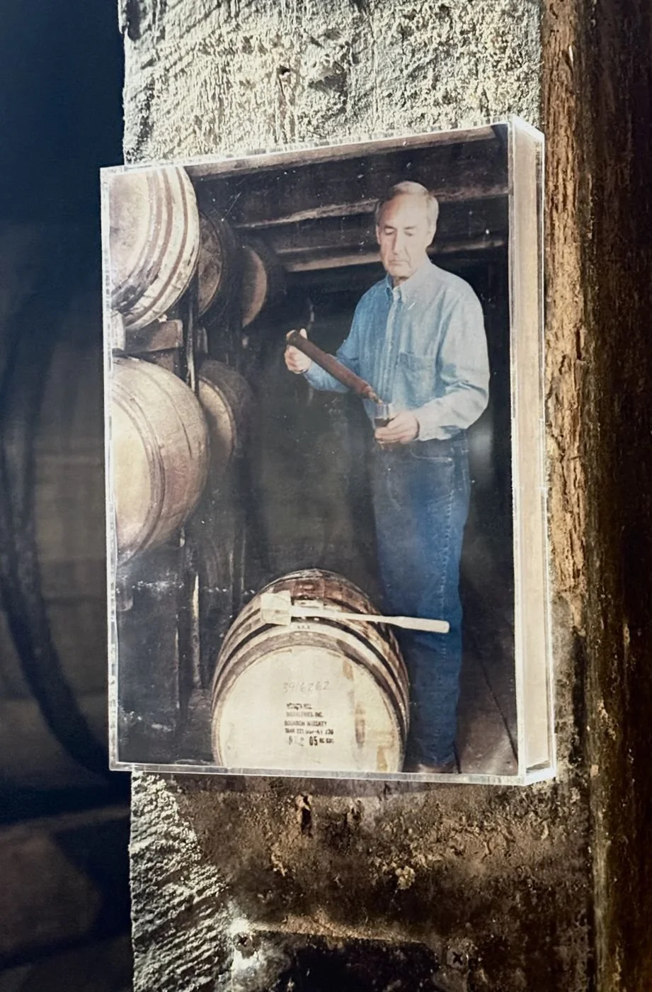 Photo of the late Parker Beam, Heaven Hill master distiller, holding a whiskey thief, displayed on a rickhouse wall.