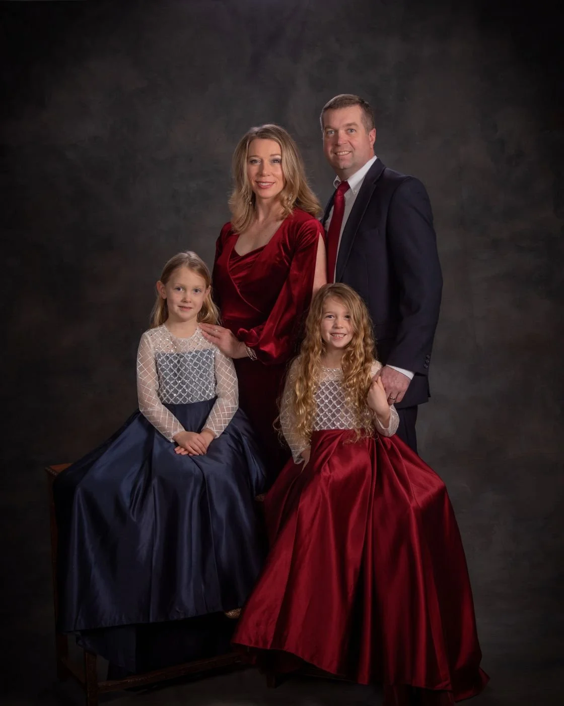 Fine art studio portrait of a family of four in coordinating burgundy and navy gowns against a dark painted backdrop
