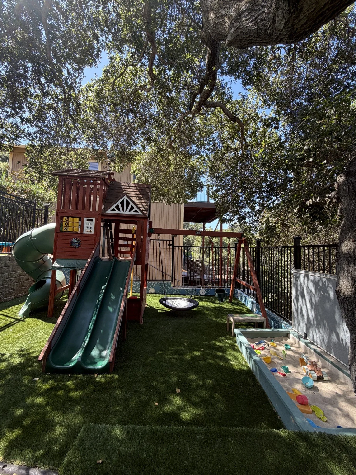 Mila-Ma’s playground with slides, swings, a sand area, and a large tree canopy