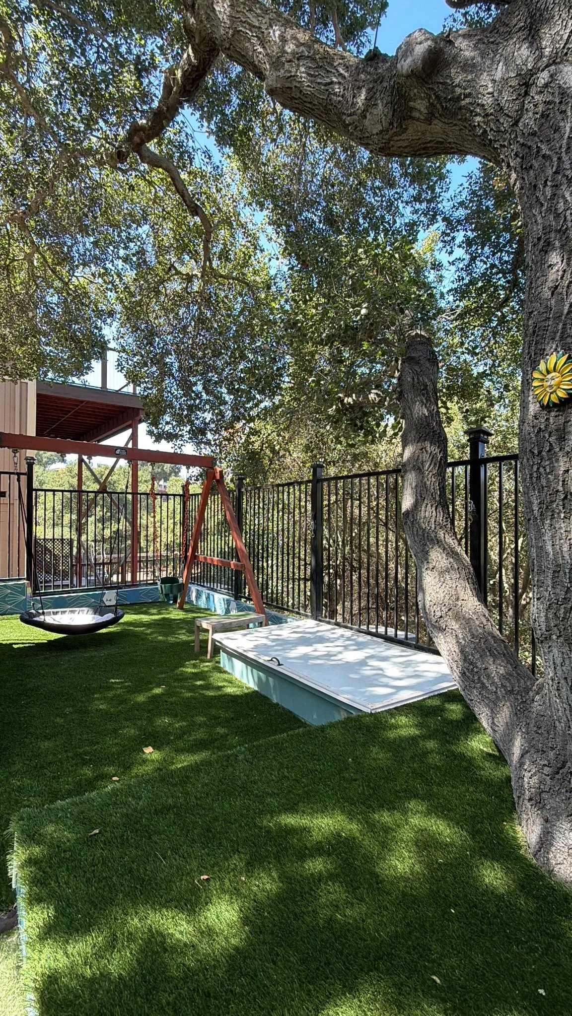 A shaded corner of the playground with a large tree, swing, and covered sand area