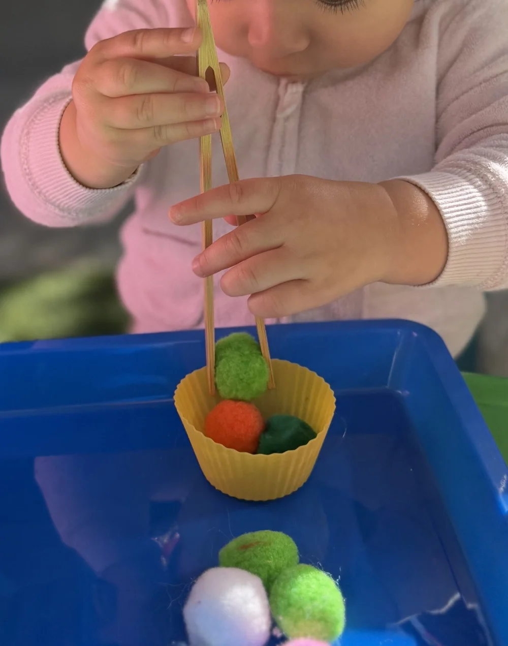 A child using wooden tongs to move colorful pompoms into a cup