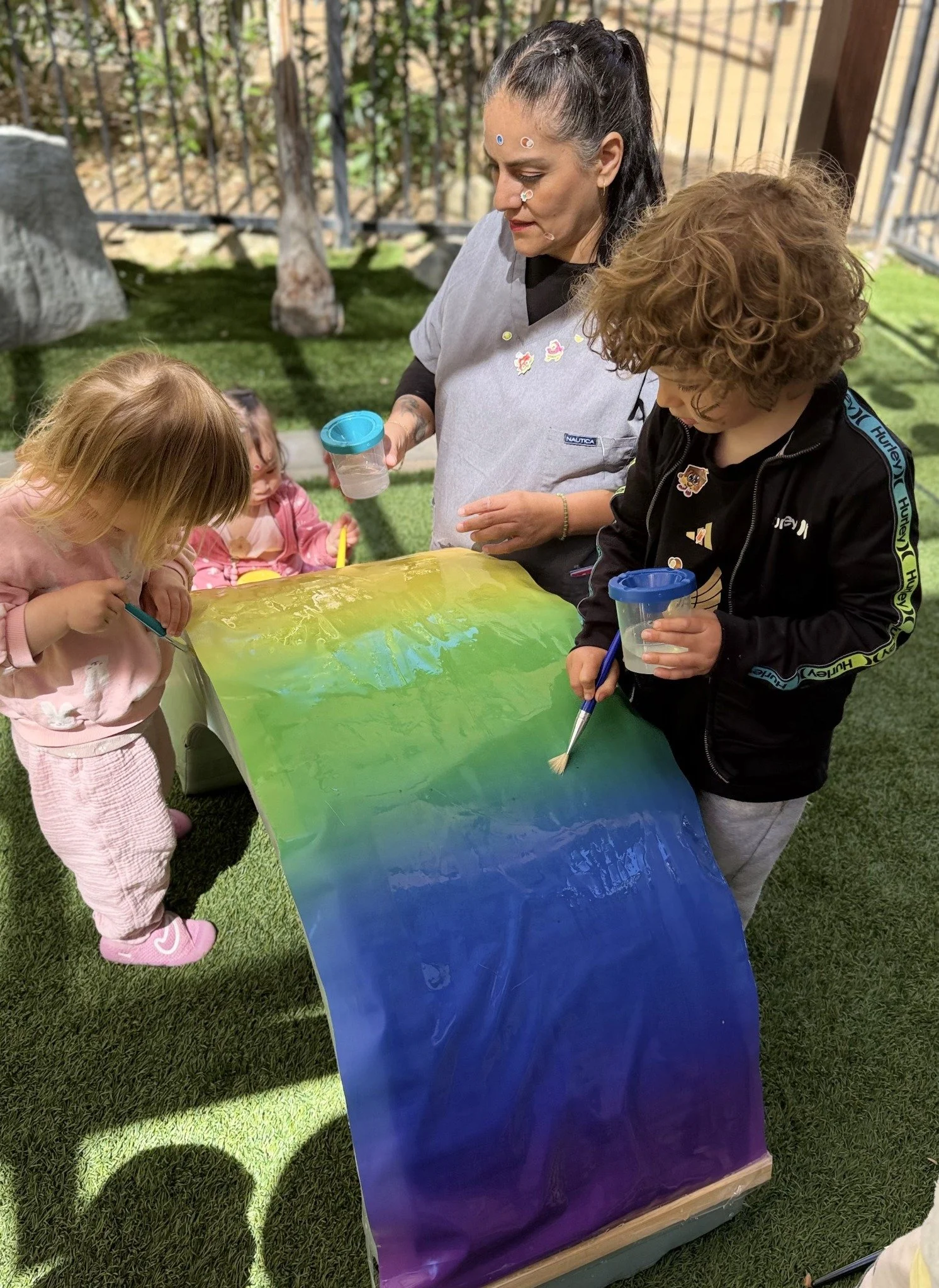 A caregiver and children painting a large colorful structure outdoors