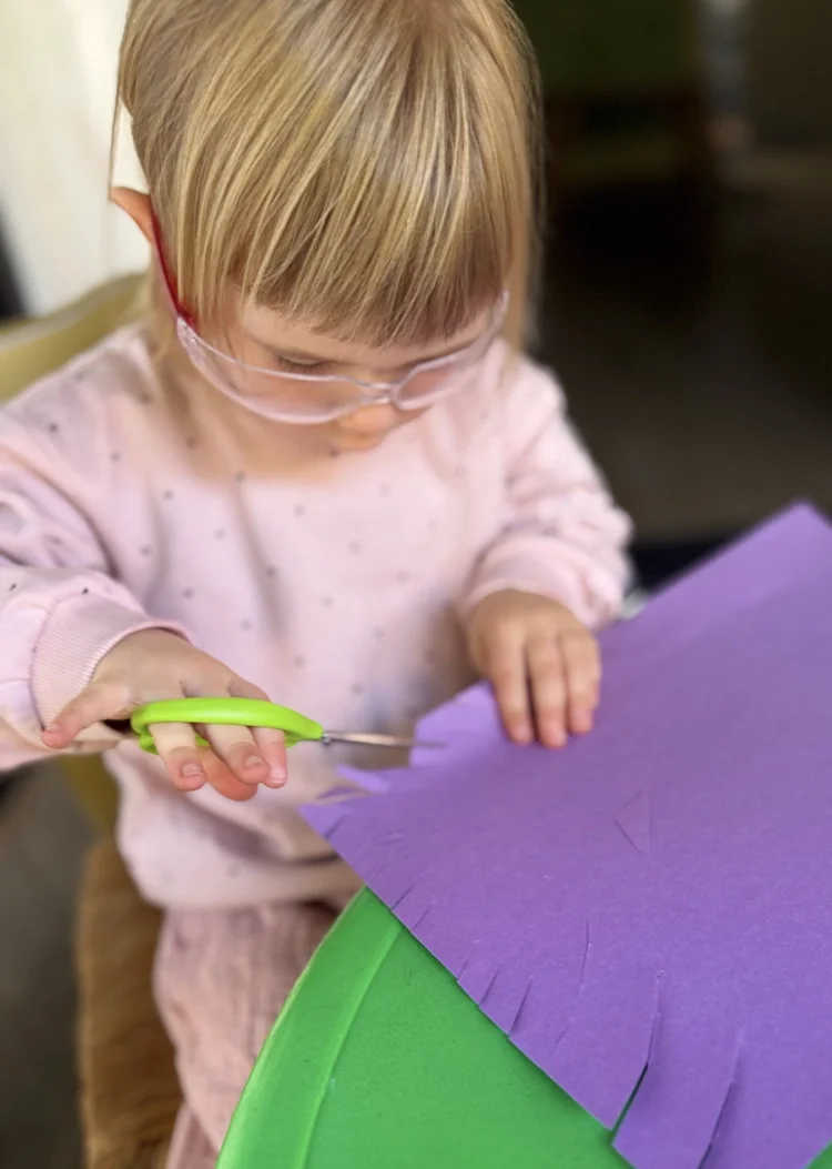 A child cutting purple paper with green scissors