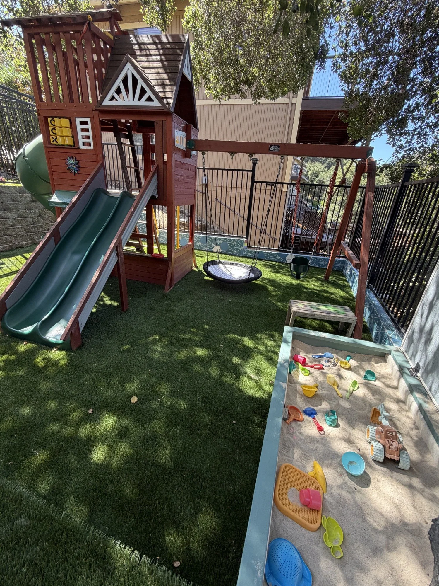 The wooden play structure, green slides, nest swing, and sand area at Mila-Ma