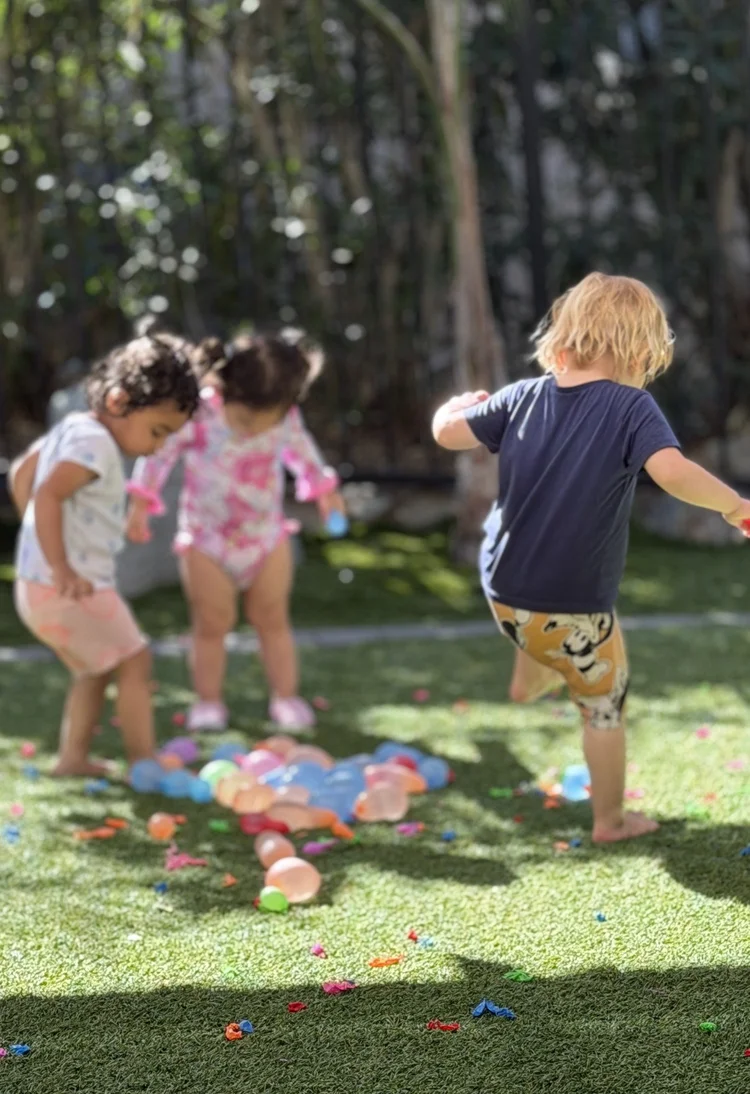 Three children exploring colorful water balloons on the grass