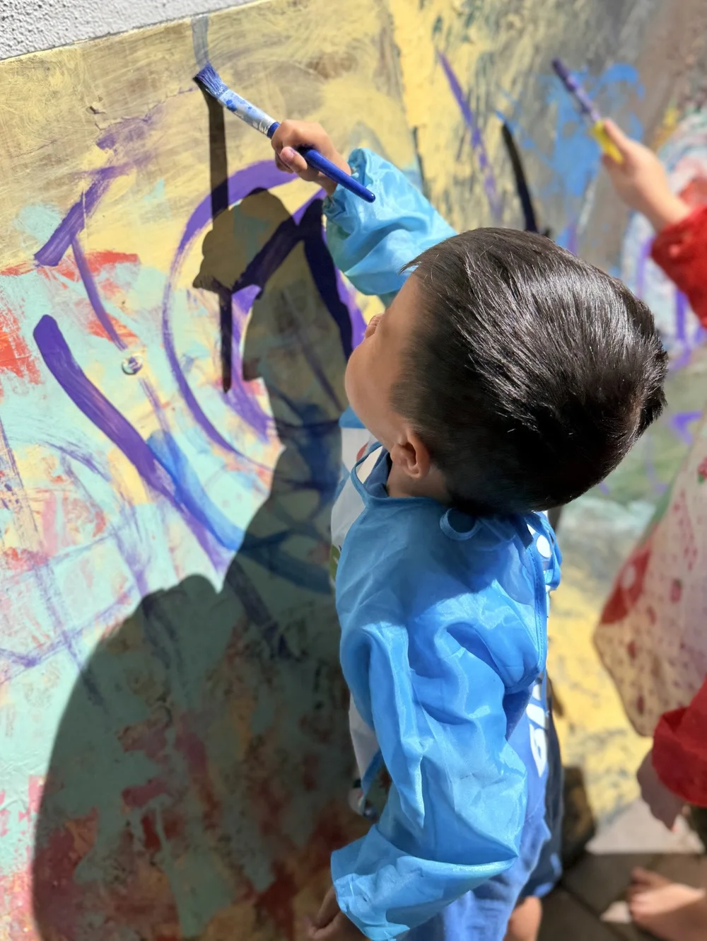 A child in a blue painting smock making bold marks on an outdoor canvas
