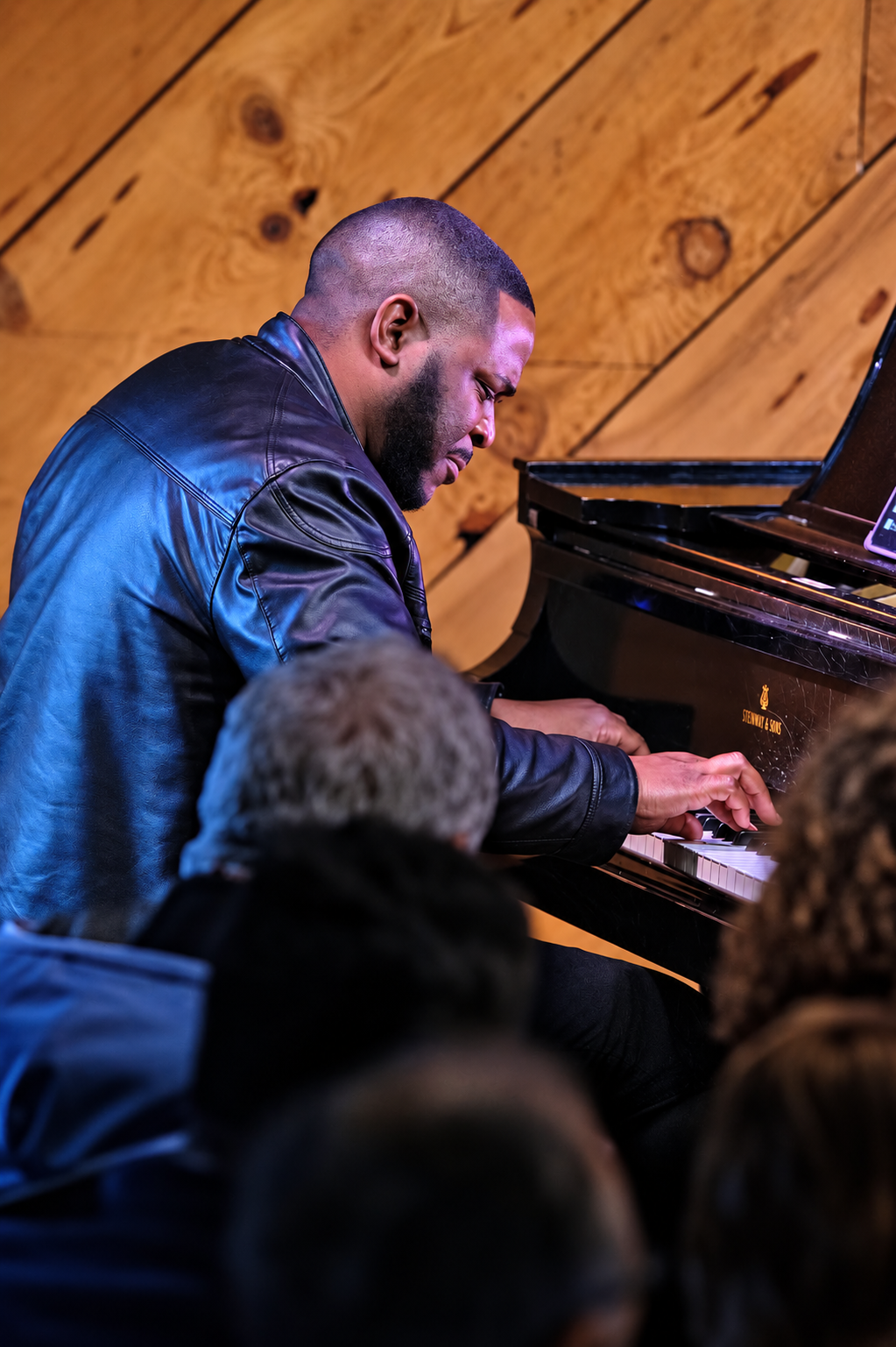 Udobi performing at a grand piano in a leather jacket, with warm wood paneling and an audience in the foreground