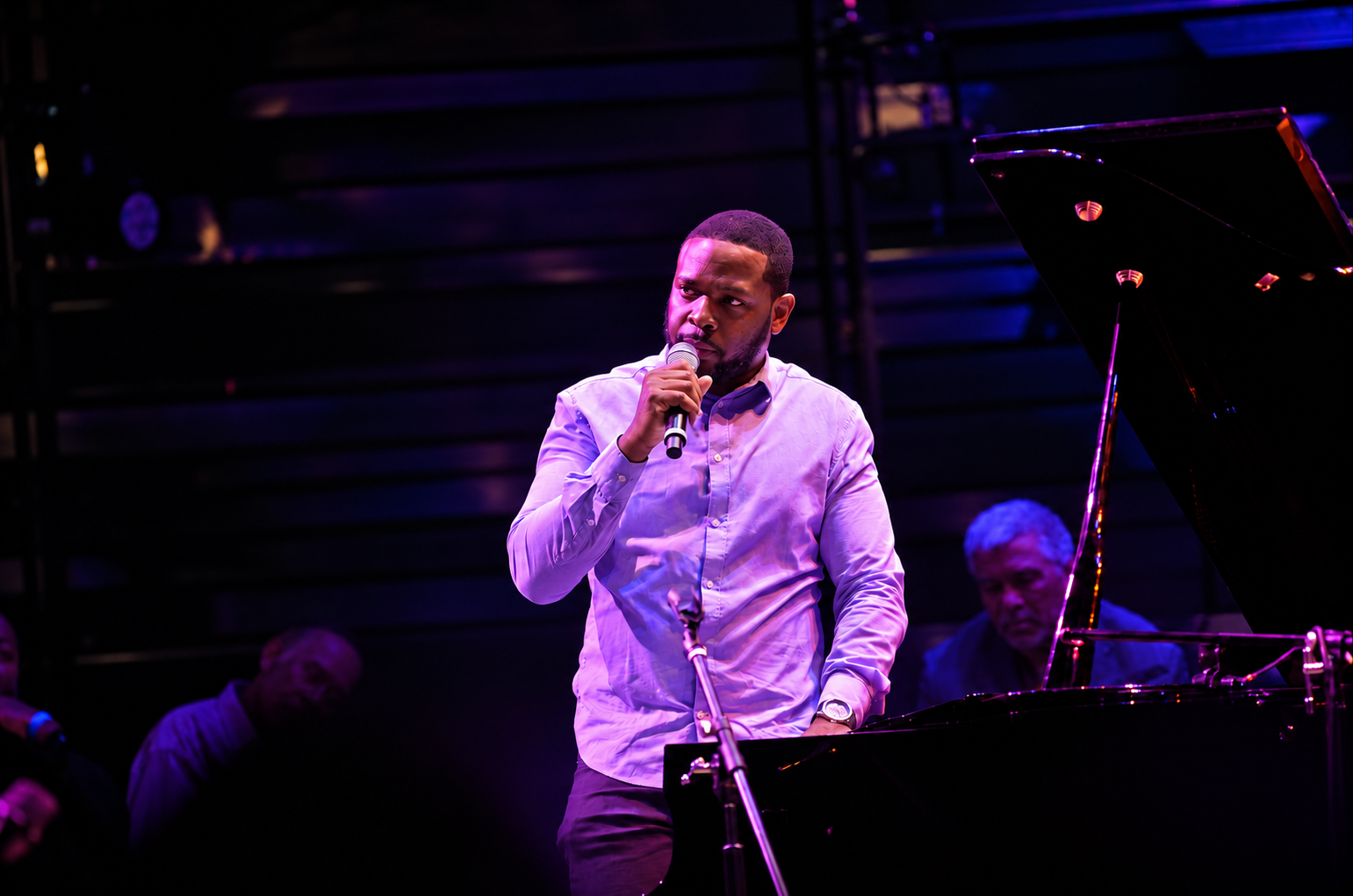 Udobi holding a microphone beside a grand piano under purple stage lights