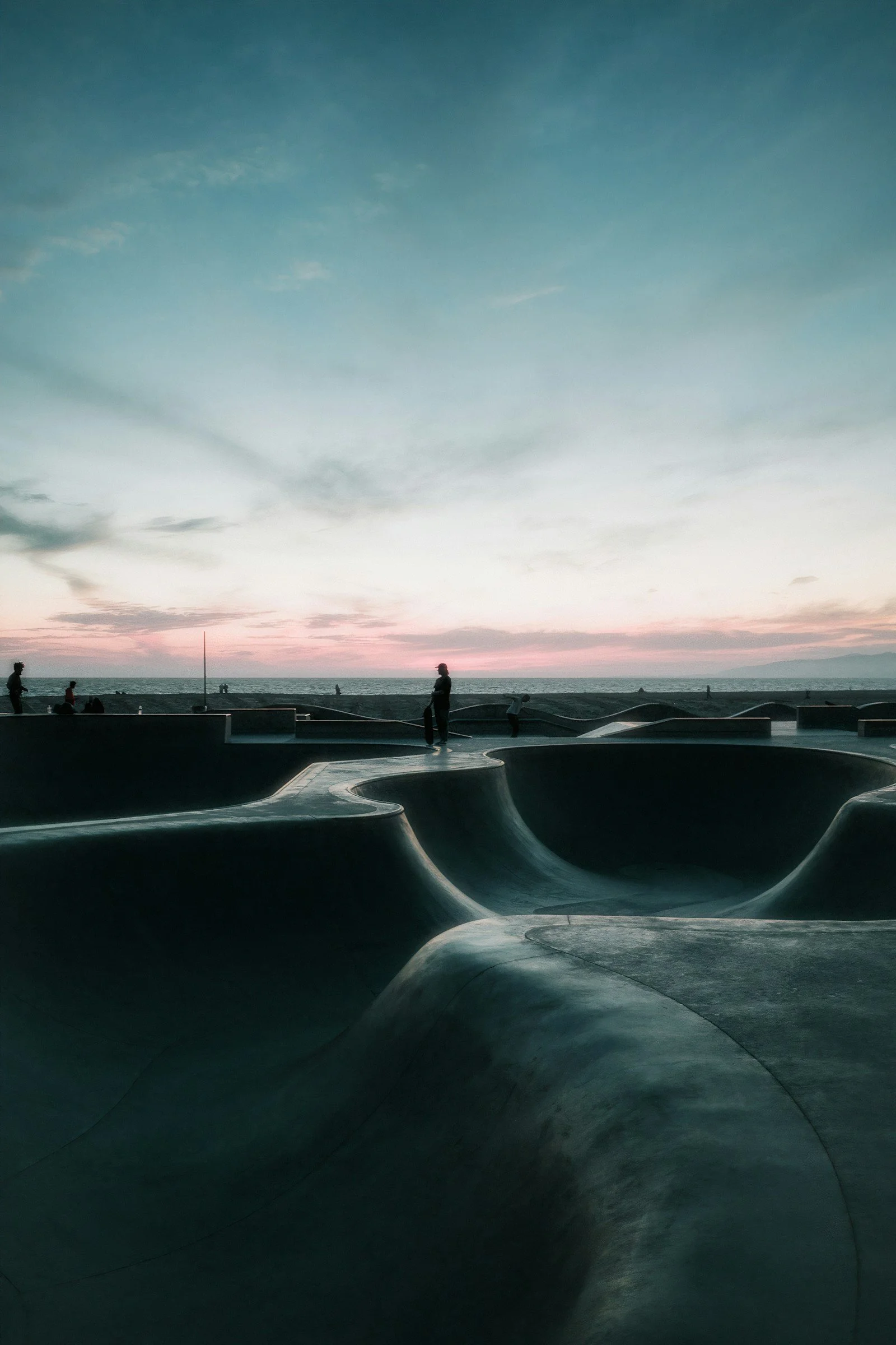 People among curved concrete skatepark bowls beside the ocean at sunset.