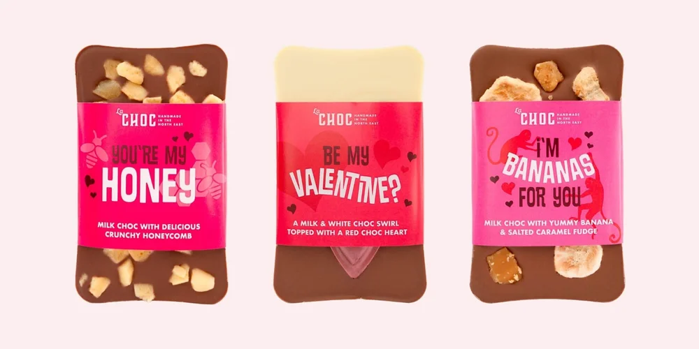 E-commerce product photography of three artisan La Chocolatrice chocolate bars featuring bright pink Valentine's Day wrappers and visible toppings like honeycomb and salted caramel fudge.