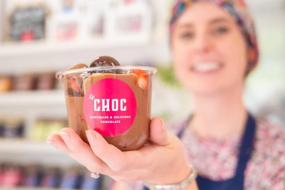 A close-up of a clear cup filled with fresh strawberries and melted chocolate featuring the pink La Choc sticker, held by a smiling staff member in the background.