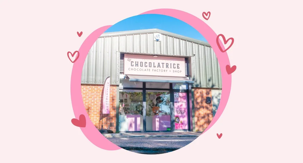 The exterior storefront of the La Chocolatrice Chocolate Factory and Shop, framed within a pink circular graphic decorated with floating heart illustrations.