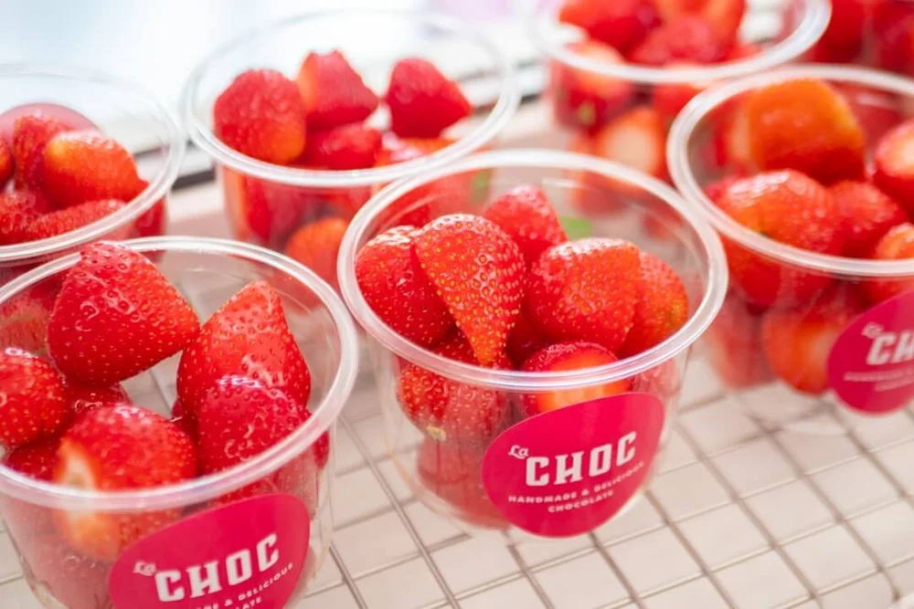 Several clear plastic cups filled with fresh red strawberries and branded with pink La Choc stickers, prepped and ready for melted chocolate.