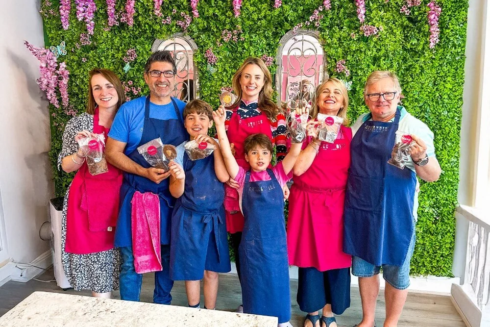A happy family wearing aprons and holding their handmade chocolate treats during a chocolate-making workshop at La Chocolatrice, posing in front of a decorative green flower wall.