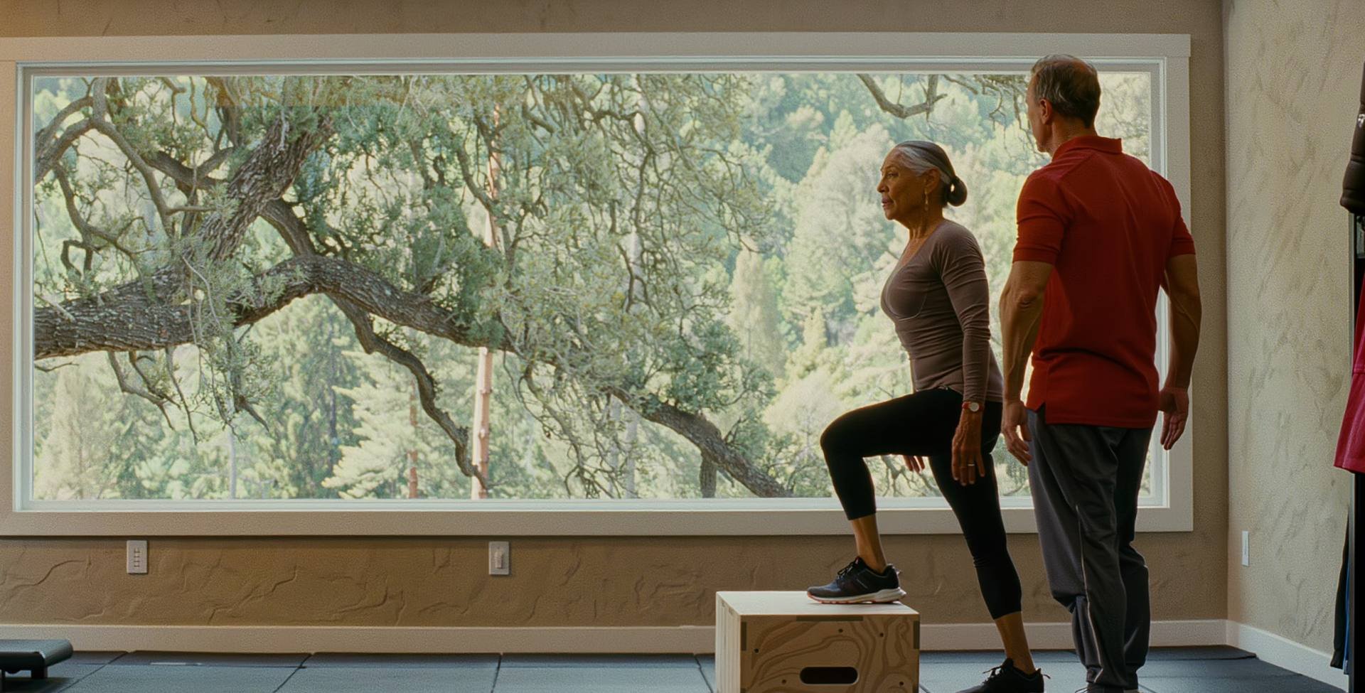 A woman in her 60s stepping up onto a box in a home gym with her coach beside her