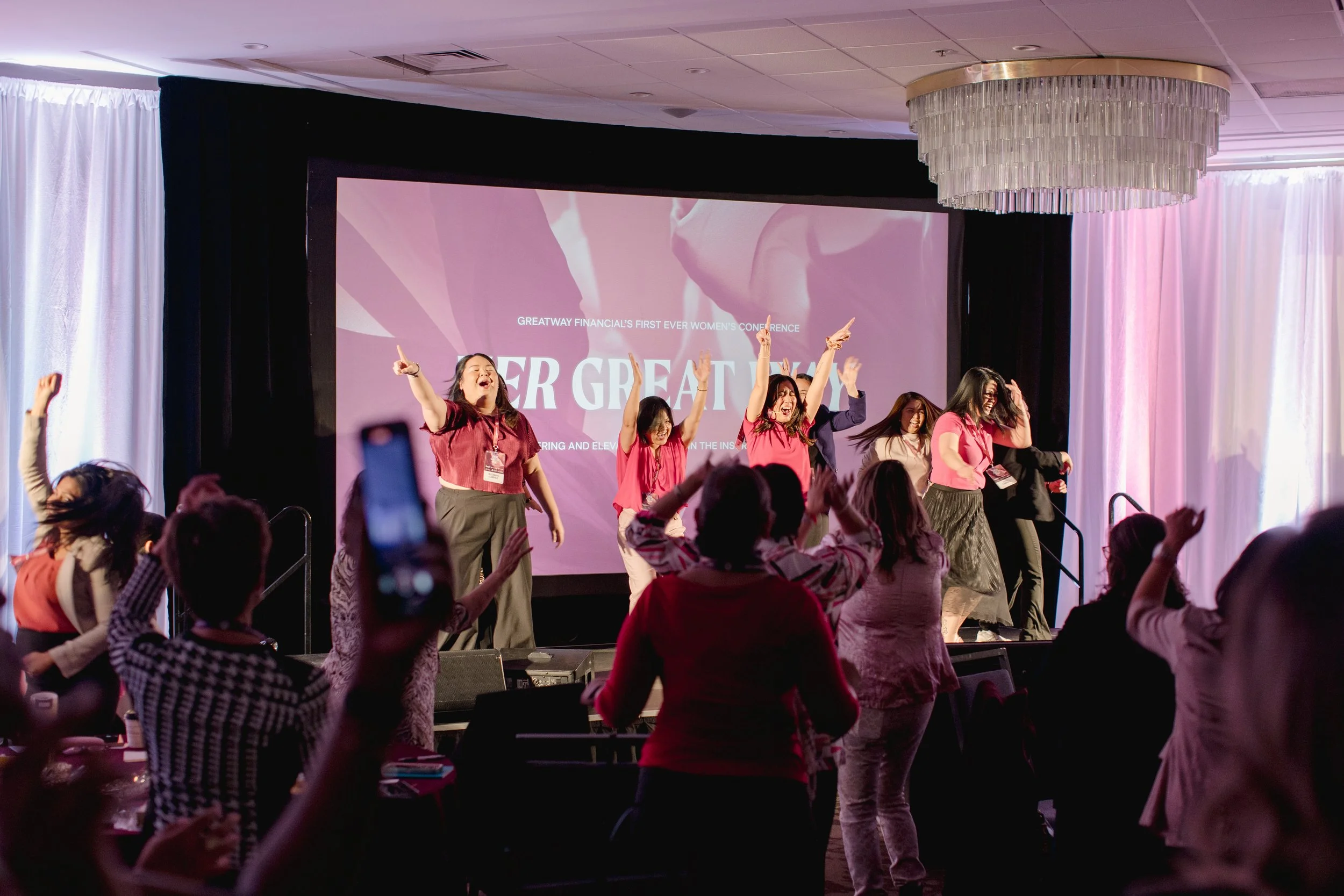 Attendees on stage at the opening of Her Greatway, hands raised in celebration.