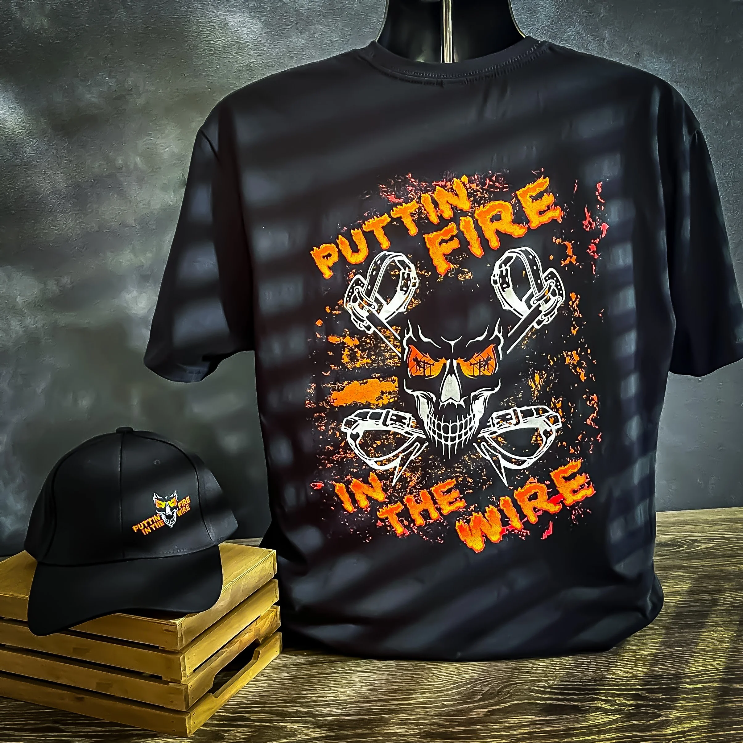 Black Putting Fire in the Wire graphic T-shirt displayed with a matching black cap on a wooden crate.