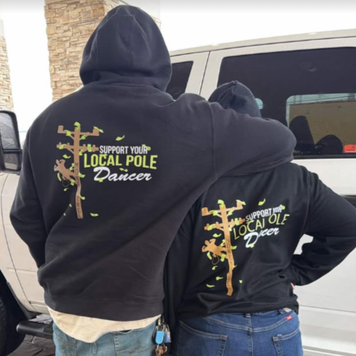 Two people photographed from behind wearing black Support Your Local Pole Dancer hoodies beside a vehicle.