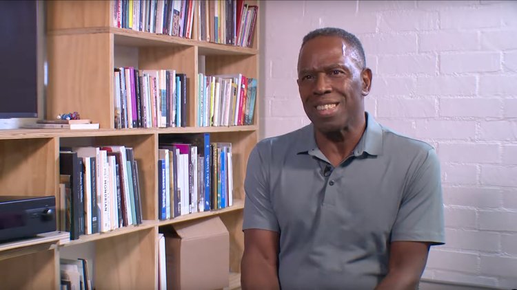 A screenshot from Charles Gaines' TAPA short documentary, with the artist smiling next to a bookshelf.