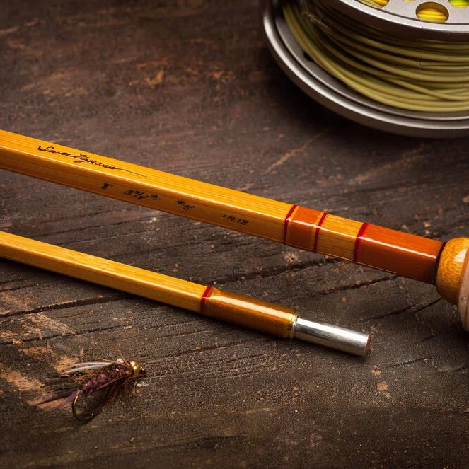 Two Sweetgrass bamboo sections on weathered wood, the upper one script-marked "Sweetgrass" with its length, weight and build date, orange wraps and a nickel-silver ferrule, a line-loaded reel and a soft-hackle fly alongside (photo: sweetgrassrods.com)