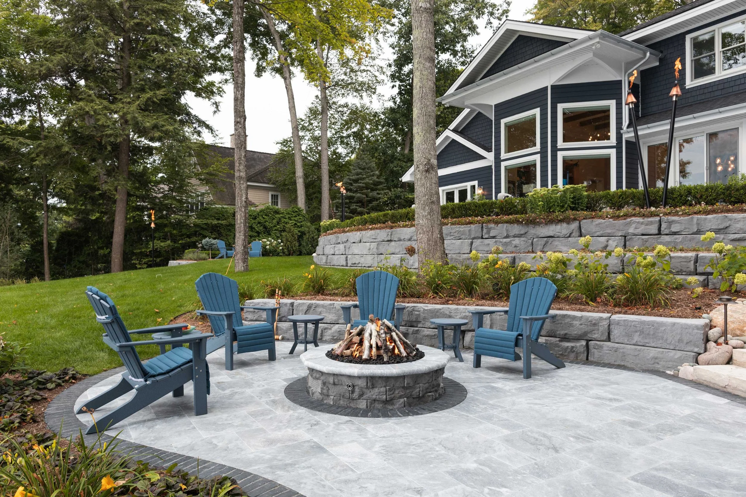Grand Ledge retaining wall backdrop behind a fire pit patio with blue Adirondack chairs and paver surface