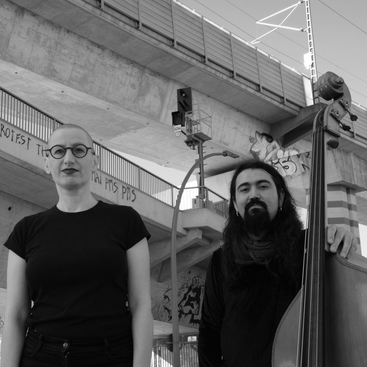 Schwarz-weiß Foto der zwei Musiker:innen unter einer Eisenbahnbrücke.