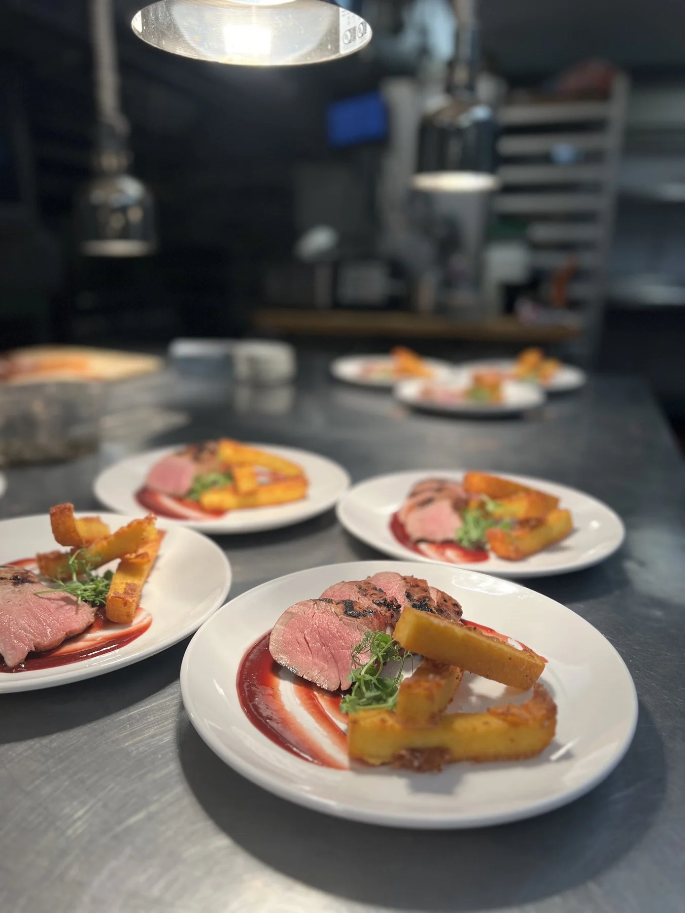 A run of plated entrées with golden batons, sauce and greens on the kitchen pass