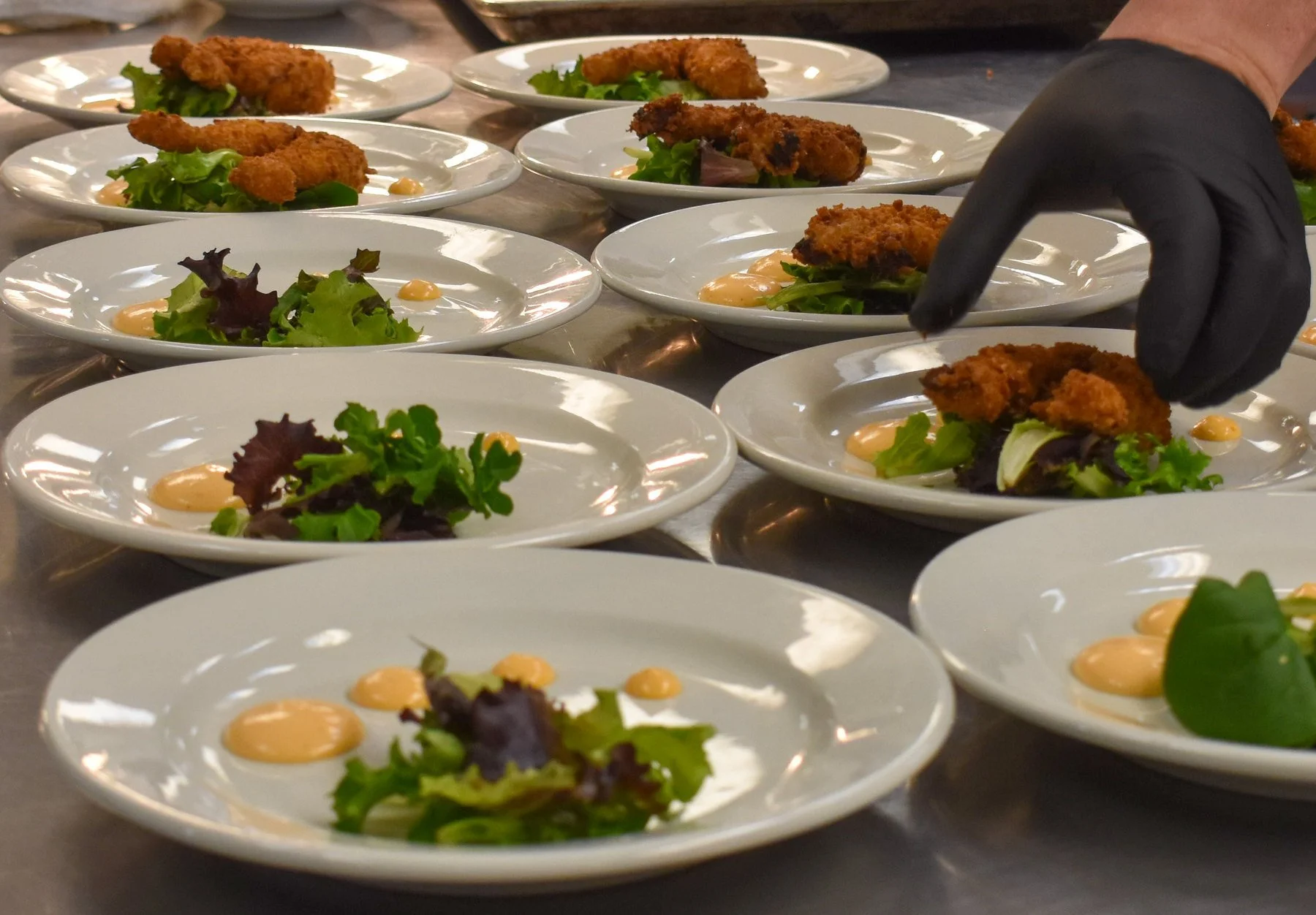A chef’s gloved hand finishing rows of appetizers with greens and sauce for service