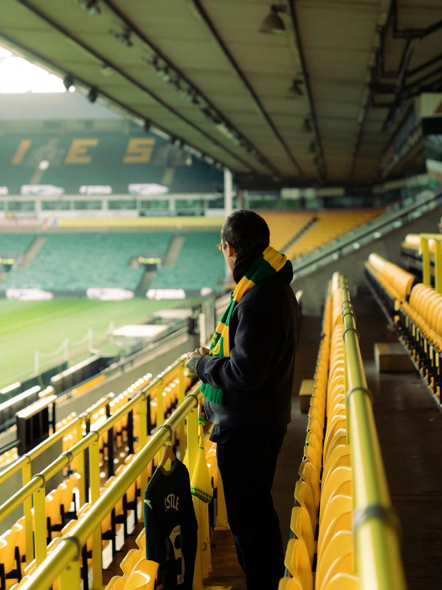 Bobby Harrison in a Norwich City scarf looking out over Carrow Road from the yellow seats, Hustle shirts on the rail