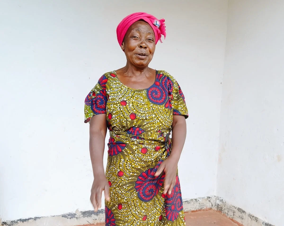 Grace, smiling, in a pink headscarf and patterned dress, standing in a plain room in Zambia.