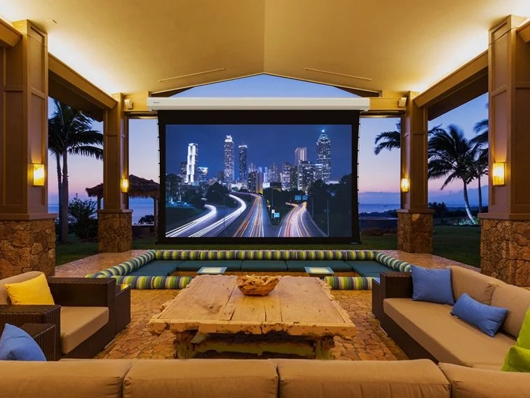 Covered outdoor living area at dusk with a Stewart Filmscreen motorized retractable projection screen lowered from a ceiling case | VIP Smart Homes