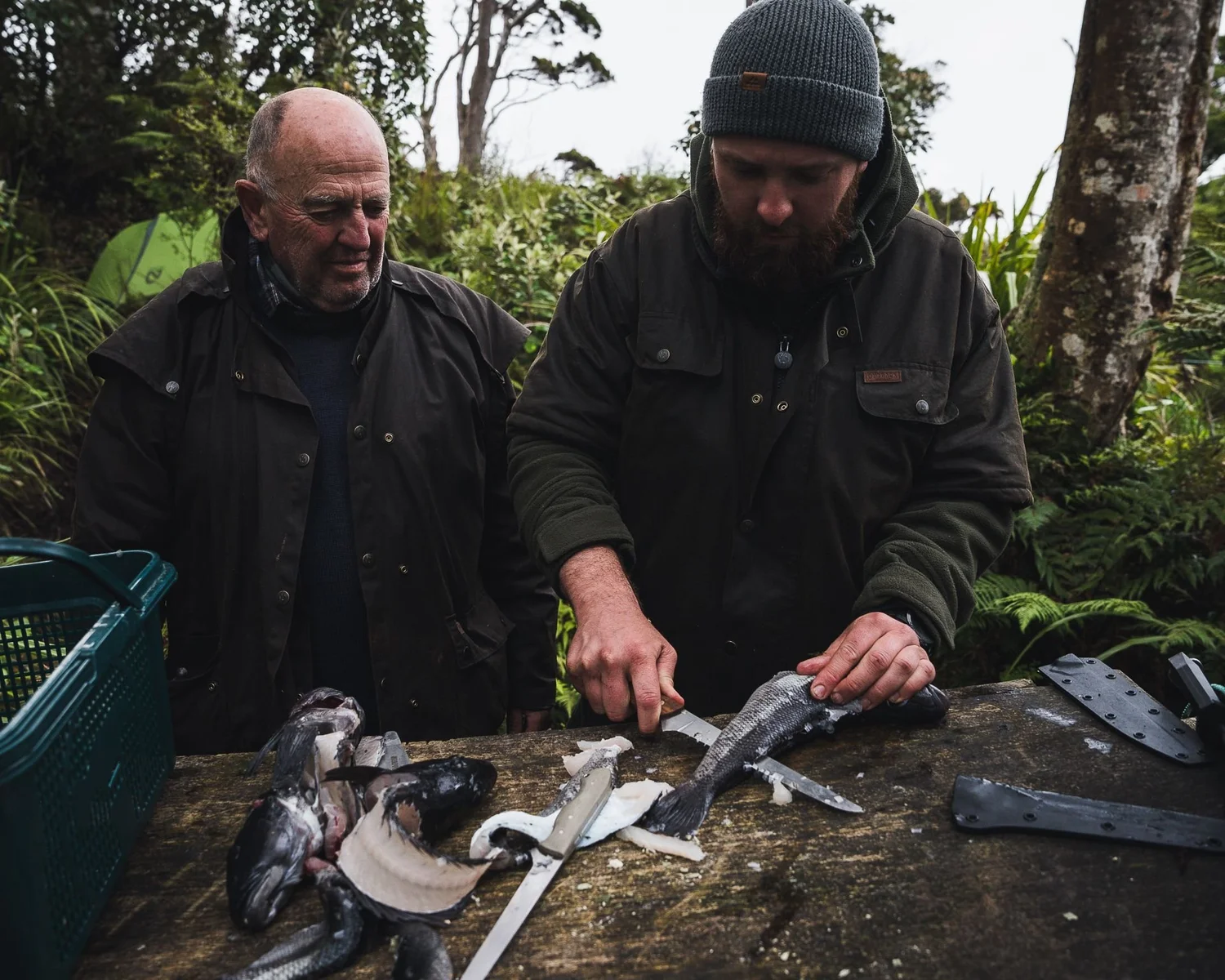 Two of the crew filleting fish with the handmade filleting knives on a log in the bush, black Kydex sheaths beside them
