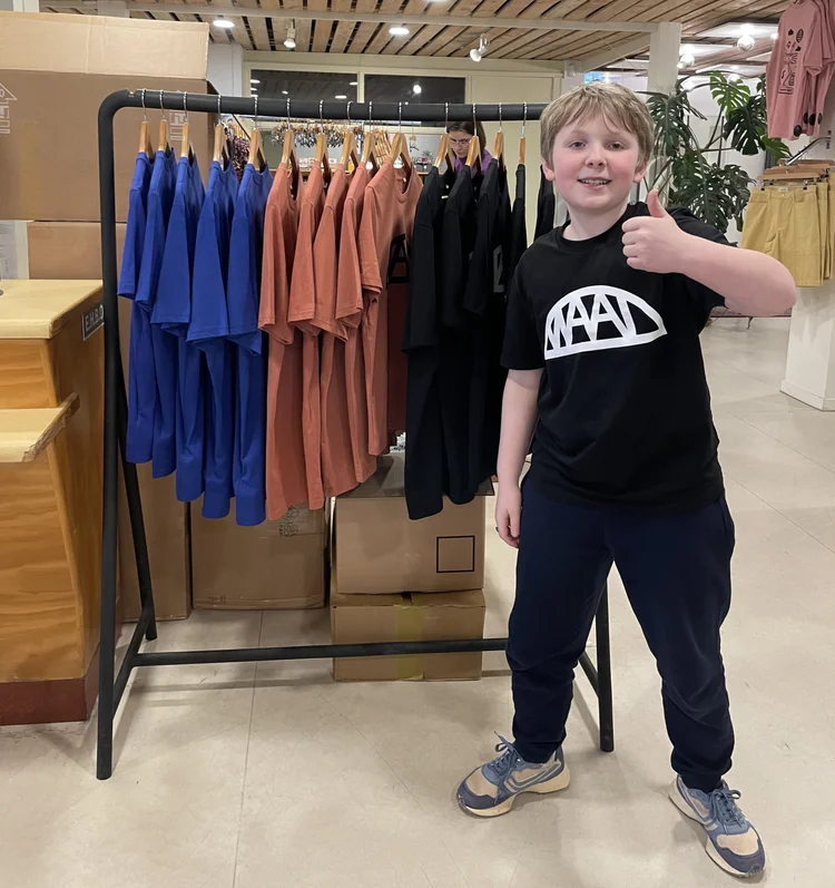 Een jongetje staat met opgestoken duim voor een kledingrek met T-shirts