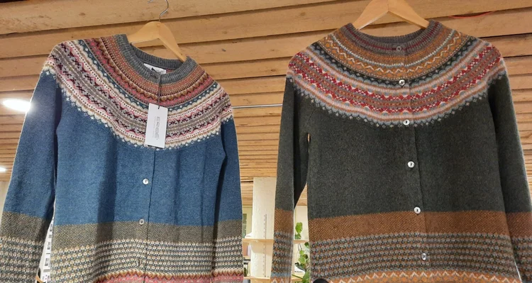 Twee vesten aan hangers, links blauw, rechts groen. Allebei hebben ze boven en onderaan ingebreide kleuren