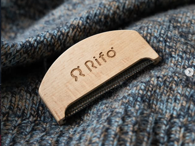 Een deel van een grijs-gemeleerde trui met daarop een houten wolkammetje met de tekst Rifo