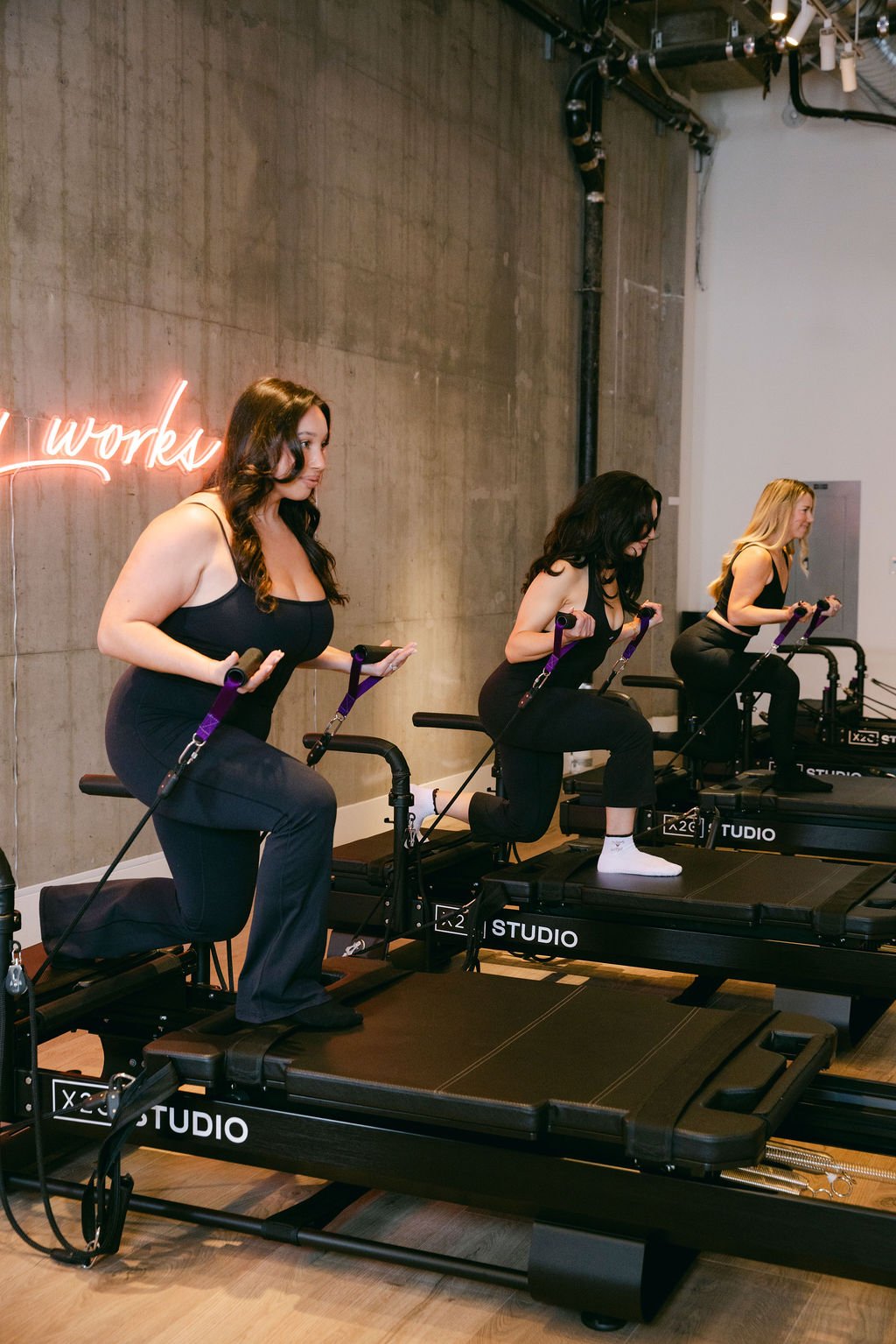 A smiling woman performing a squat exercise on the XFormer machine at x2o Studio, with other members working out in the background in a modern studio with concrete walls
