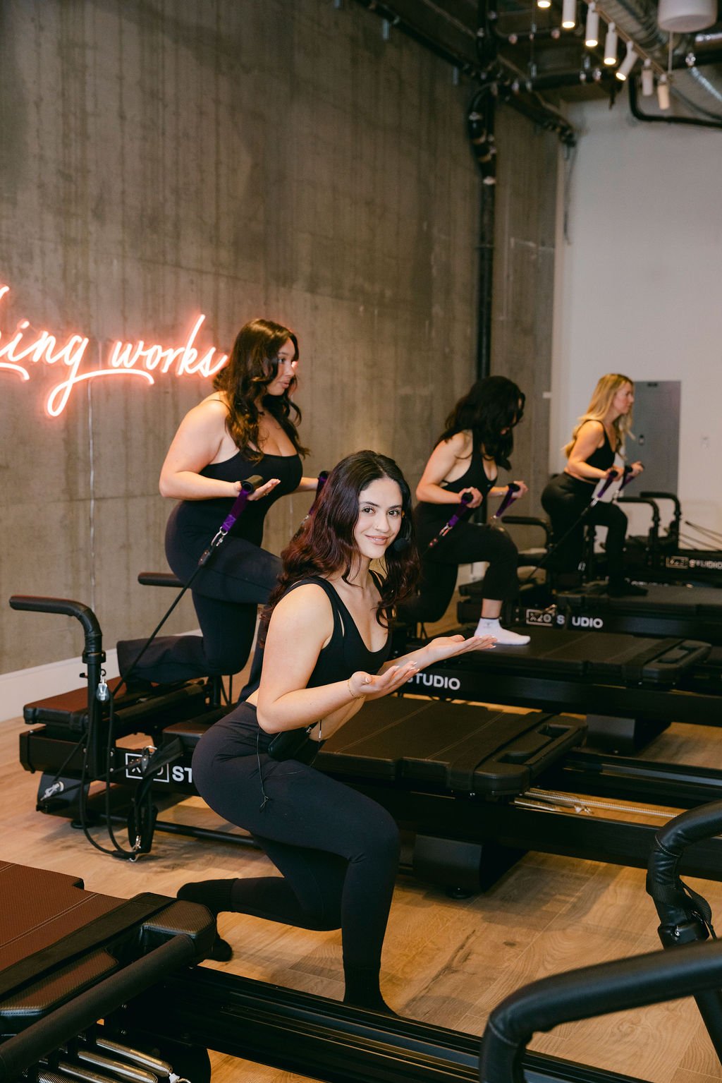 A smiling woman doing a deep squat on the XFormer machine at x2o Studio Fremont while other members work out behind her, all dressed in black athletic wear in a modern studio with a neon sign on the concrete wall
