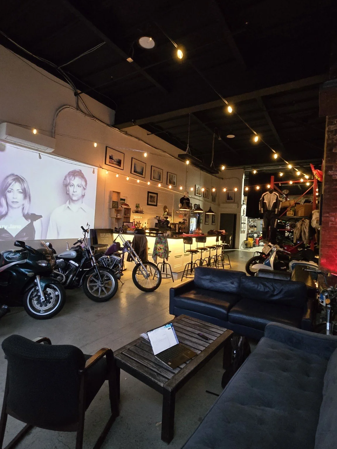 Displaced clubhouse with sofas, warm bar lighting, motorcycles and projection screen
