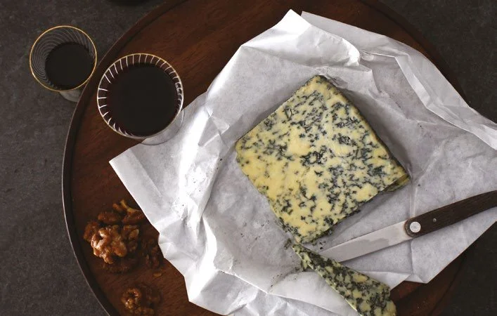 A wedge of blue cheese on wax paper with walnuts and two glasses of red wine