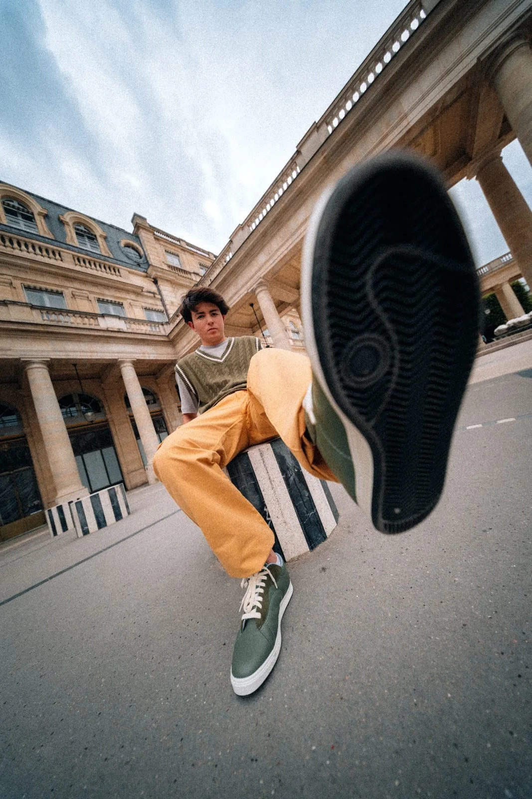 Jeune homme assis au Palais-Royal tendant la semelle de sa sneaker Twins for Peace vers l'objectif